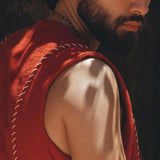 A man with a beard and an earring looks over his shoulder, wearing an AYA Sacred Wear Red Hand Embroidered Vest for Men. The lighting casts shadows on his skin, highlighting his muscular arm and intense gaze.