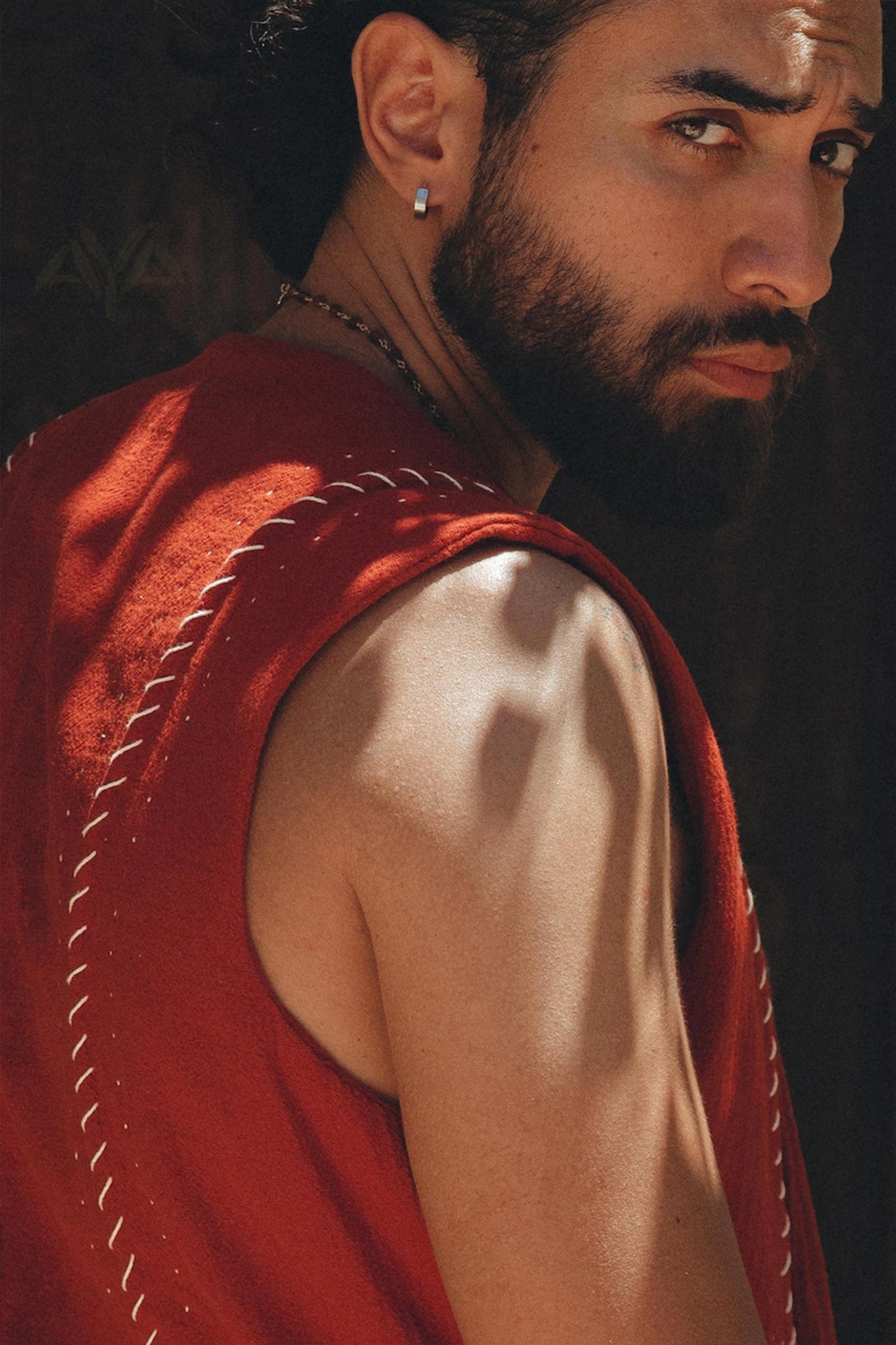 A man with a beard and an earring looks over his shoulder, wearing an AYA Sacred Wear Red Hand Embroidered Vest for Men. The lighting casts shadows on his skin, highlighting his muscular arm and intense gaze.