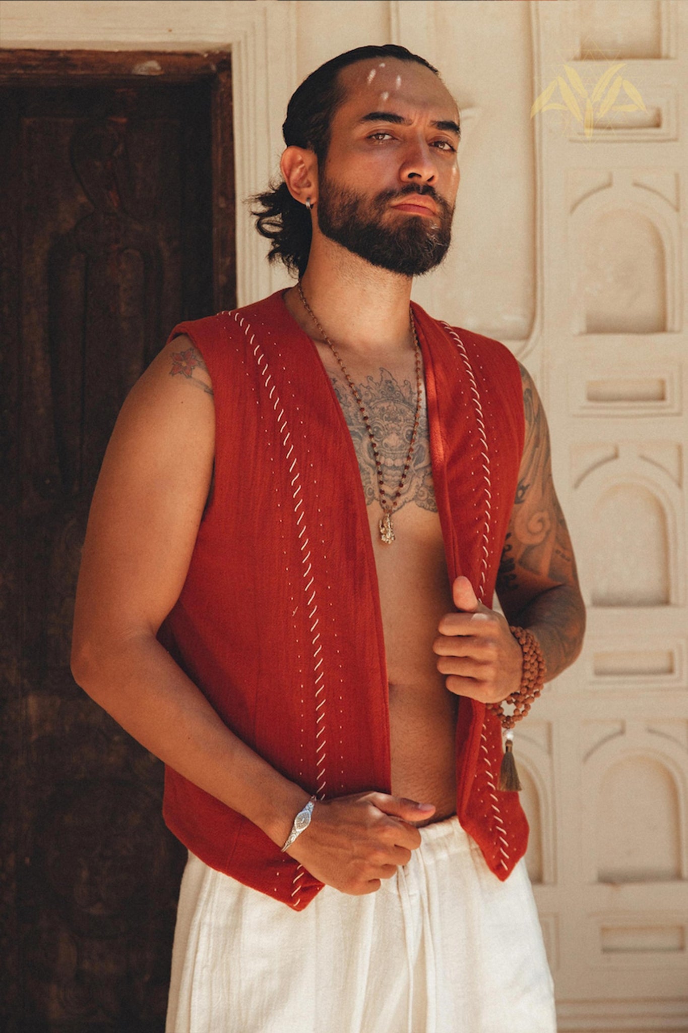 A man with a beard stands confidently in front of a carved wooden door, wearing an AYA Sacred Wear Red Hand Embroidered Vest for Men and white pants, showcasing necklaces and tattoos.