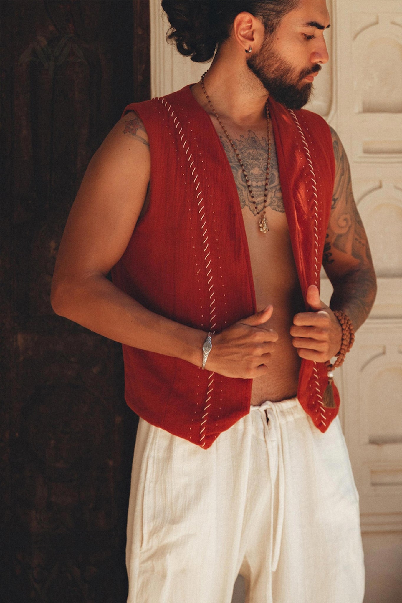 A man with a beard stands shirtless, adorned in a Red Hand Embroidered Vest for Men by AYA Sacred Wear and white pants. He accessorizes with multiple necklaces and a bracelet, posing against a decorative wall with his gaze directed to the side.