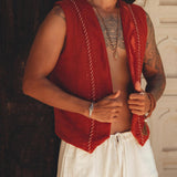 A man with a beard stands shirtless, adorned in a Red Hand Embroidered Vest for Men by AYA Sacred Wear and white pants. He accessorizes with multiple necklaces and a bracelet, posing against a decorative wall with his gaze directed to the side.