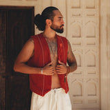 A man with a beard and a bun poses in front of an ornate wall, donning the Red Hand Embroidered Vest for Men from AYA Sacred Wear. The sleeveless vest, crafted from organic cotton, is paired with white pants. One hand grasps the vest while the other rests by his side, accentuating his necklaces and tattoos.