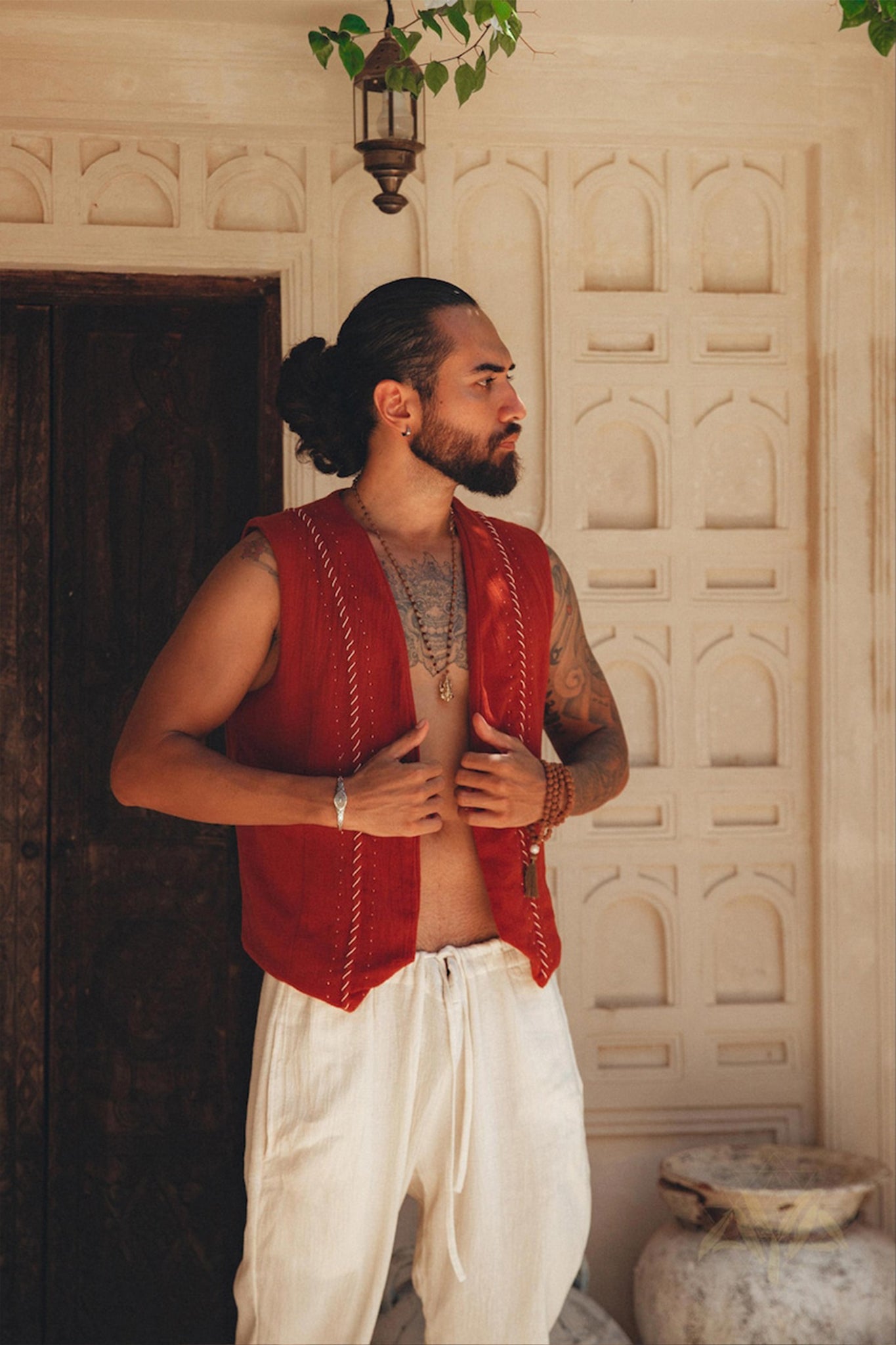 A man with a beard and a bun poses in front of an ornate wall, donning the Red Hand Embroidered Vest for Men from AYA Sacred Wear. The sleeveless vest, crafted from organic cotton, is paired with white pants. One hand grasps the vest while the other rests by his side, accentuating his necklaces and tattoos.