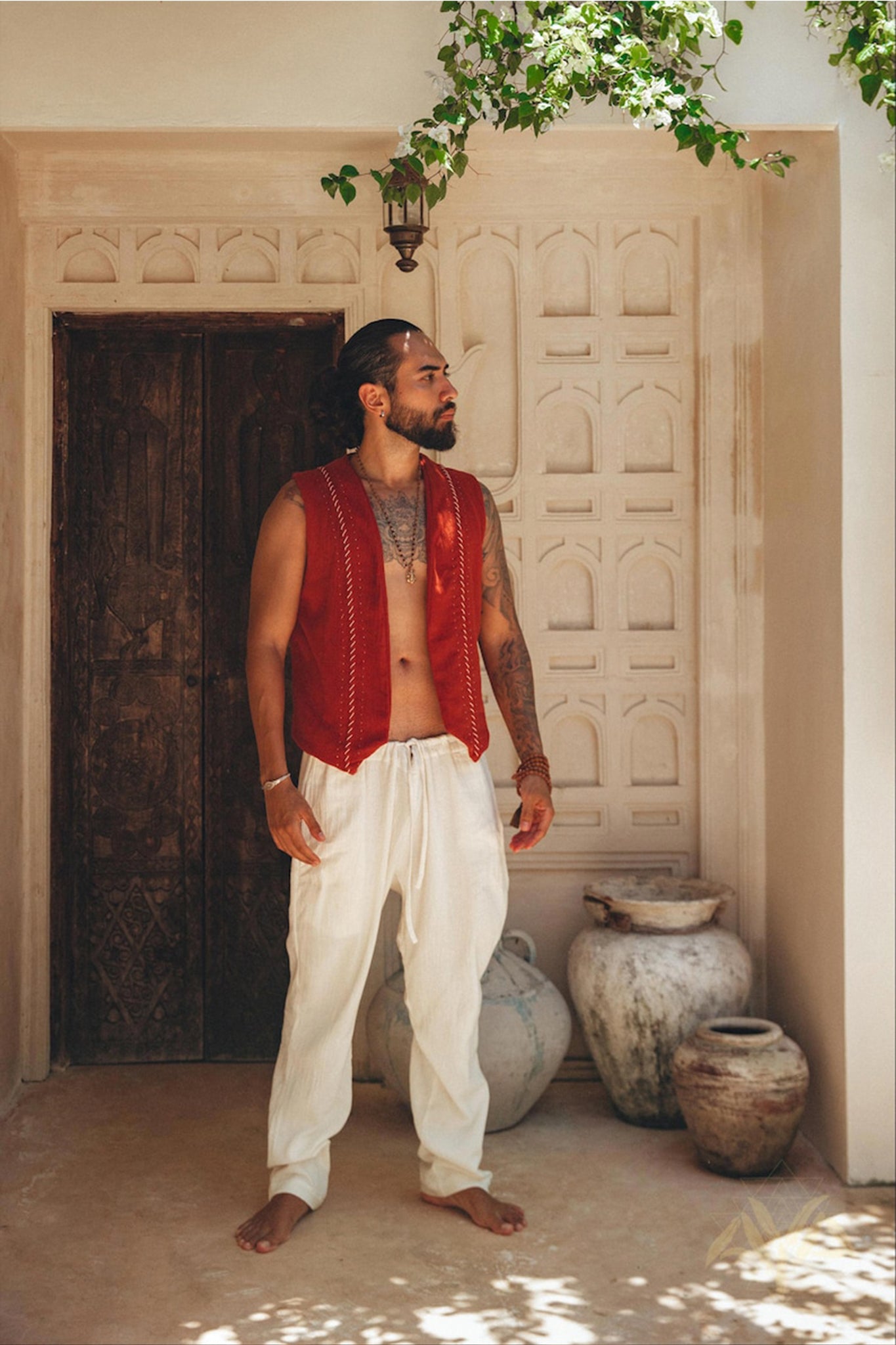 A man stands in front of an ornate wooden door, clad in a vibrant Red Hand Embroidered Vest by AYA Sacred Wear and white pants. He is barefoot and surrounded by earthen pots, as sunlight filters through the leaves, creating intricate patterns on the wall.