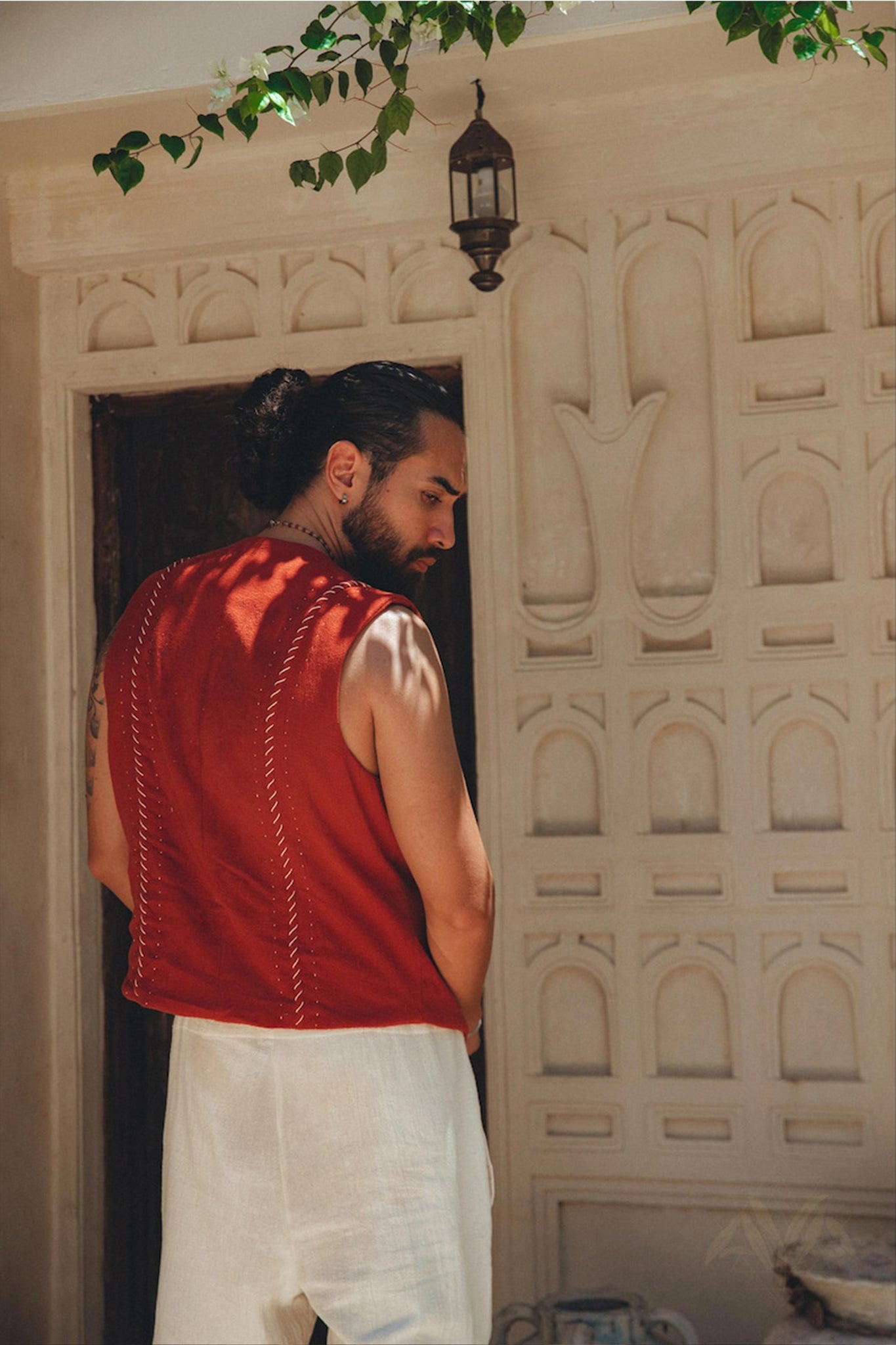 A person with long hair tied in a bun stands with their back to the camera, wearing a Red Hand Embroidered Vest for Men by AYA Sacred Wear and light-colored pants. They are near a textured wall adorned with plants and a lantern.