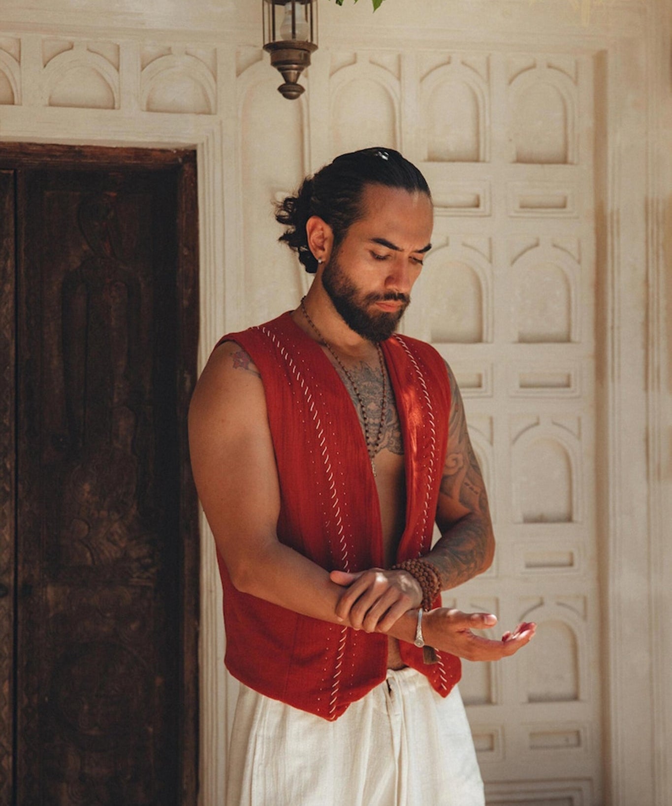 A person with long hair tied back stands by an ornate door, wearing the Red Hand Embroidered Vest for Men from AYA Sacred Wear and white pants. They are looking down, adjusting a wrist bracelet. The background features intricate stonework and a small hanging lantern.