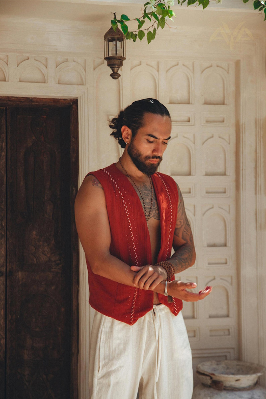 A person with long hair tied back stands by an ornate door, wearing the Red Hand Embroidered Vest for Men from AYA Sacred Wear and white pants. They are looking down, adjusting a wrist bracelet. The background features intricate stonework and a small hanging lantern.