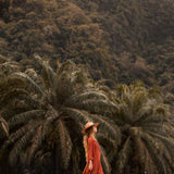 In a field of tall grass, a woman in AYA Sacred Wear's Red Kannika Shirt Dress and wide-brimmed hat gazes upward. The lush green palms and forested hillside behind evoking serenity, amplifying the tranquility brought by her organic cotton dress dyed with botanical colors.