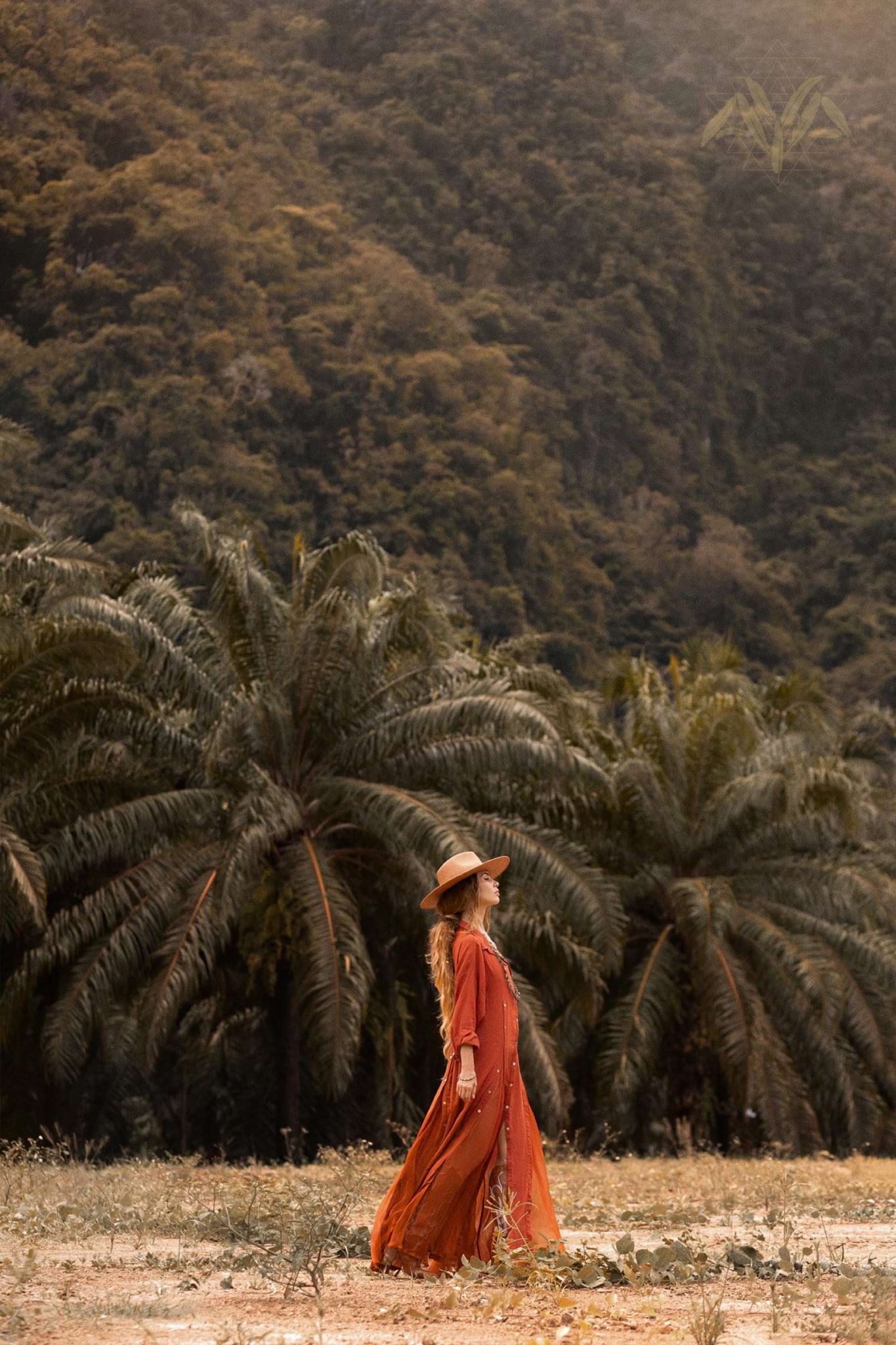 In a field of tall grass, a woman in AYA Sacred Wear's Red Kannika Shirt Dress and wide-brimmed hat gazes upward. The lush green palms and forested hillside behind evoking serenity, amplifying the tranquility brought by her organic cotton dress dyed with botanical colors.