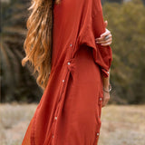 In a serene outdoor setting, an individual with long, wavy hair wears AYA Sacred Wear's Red Kannika Shirt Dress, made from lightweight fabric and botanical dyes. The dress features a multiway buttoned back design against blurred greenery.