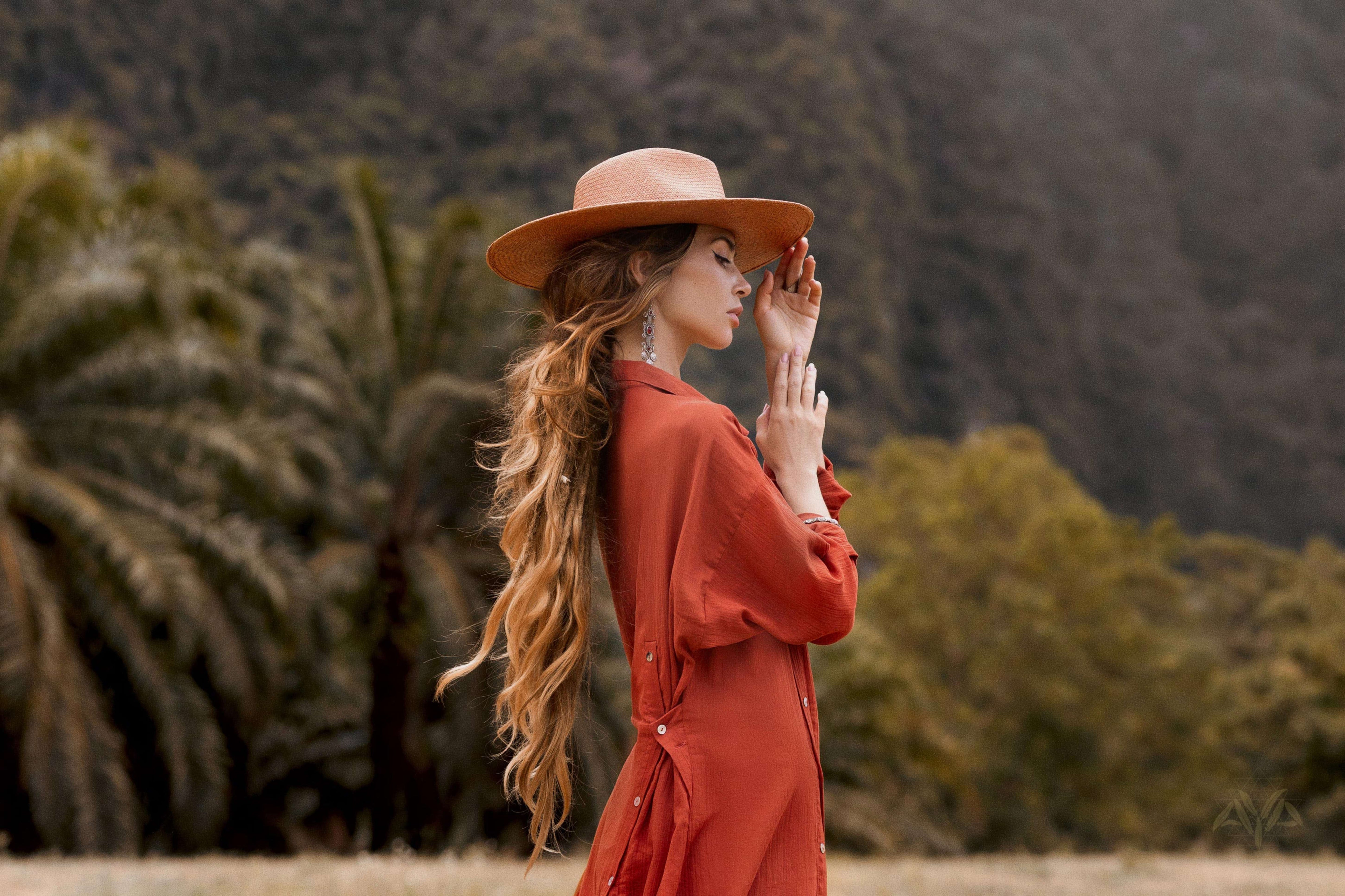 A woman with long hair stands in profile, wearing a wide-brimmed hat and the Red Kannika Shirt Dress by AYA Sacred Wear. Crafted from organic cotton and tinted with botanical dyes, she holds her hand near her face as sunlight highlights her amidst trees and mountain scenery.