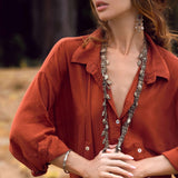 A woman outdoors in an orange button-up dress, similar to AYA Sacred Wear's Red Kannika Shirt Dress, with a wide-brimmed straw hat. She's accessorized with long necklaces, intricate earrings, and a bracelet. Her gaze is directed sideways against a backdrop of blurred foliage.