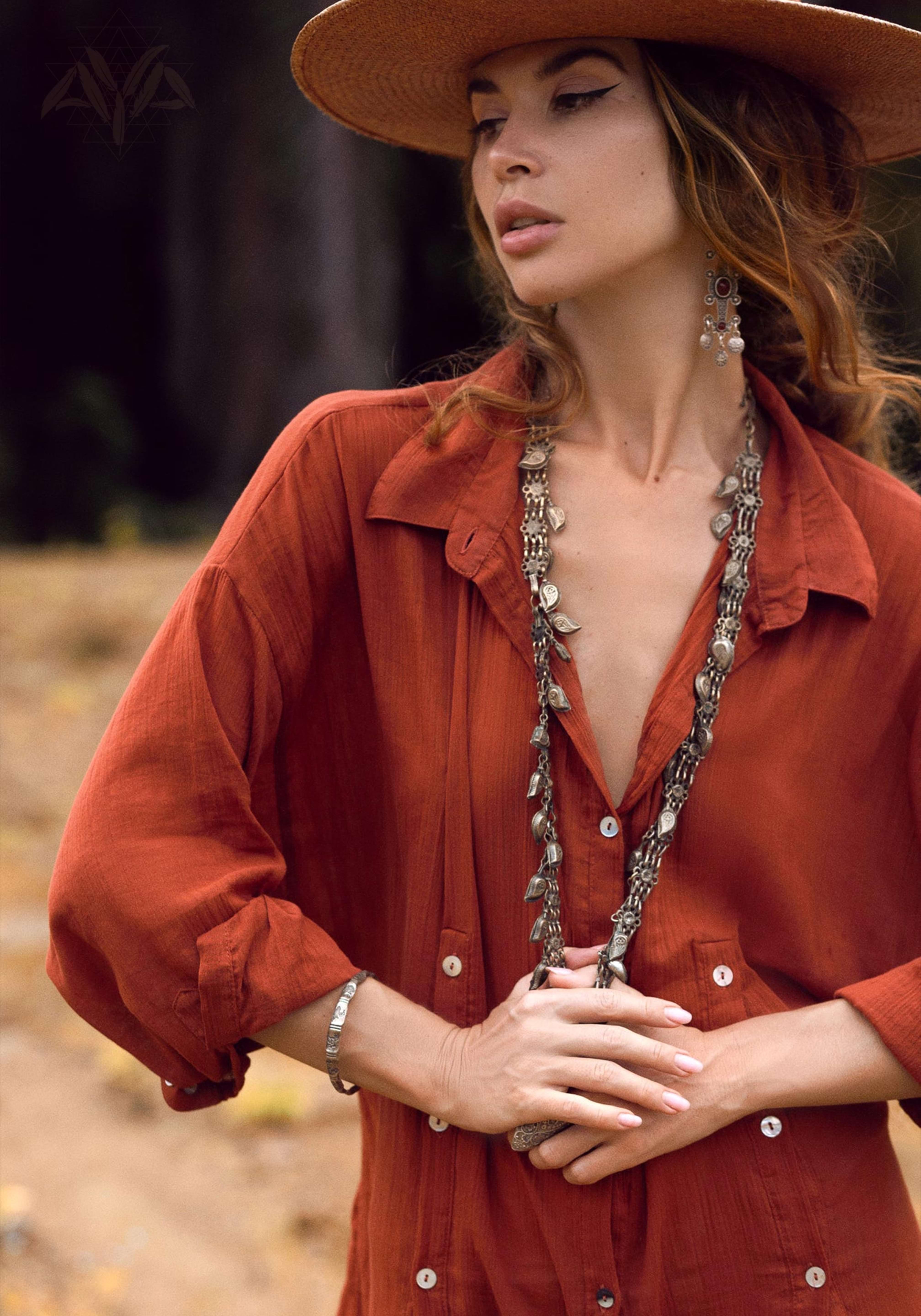 A woman outdoors in an orange button-up dress, similar to AYA Sacred Wear's Red Kannika Shirt Dress, with a wide-brimmed straw hat. She's accessorized with long necklaces, intricate earrings, and a bracelet. Her gaze is directed sideways against a backdrop of blurred foliage.