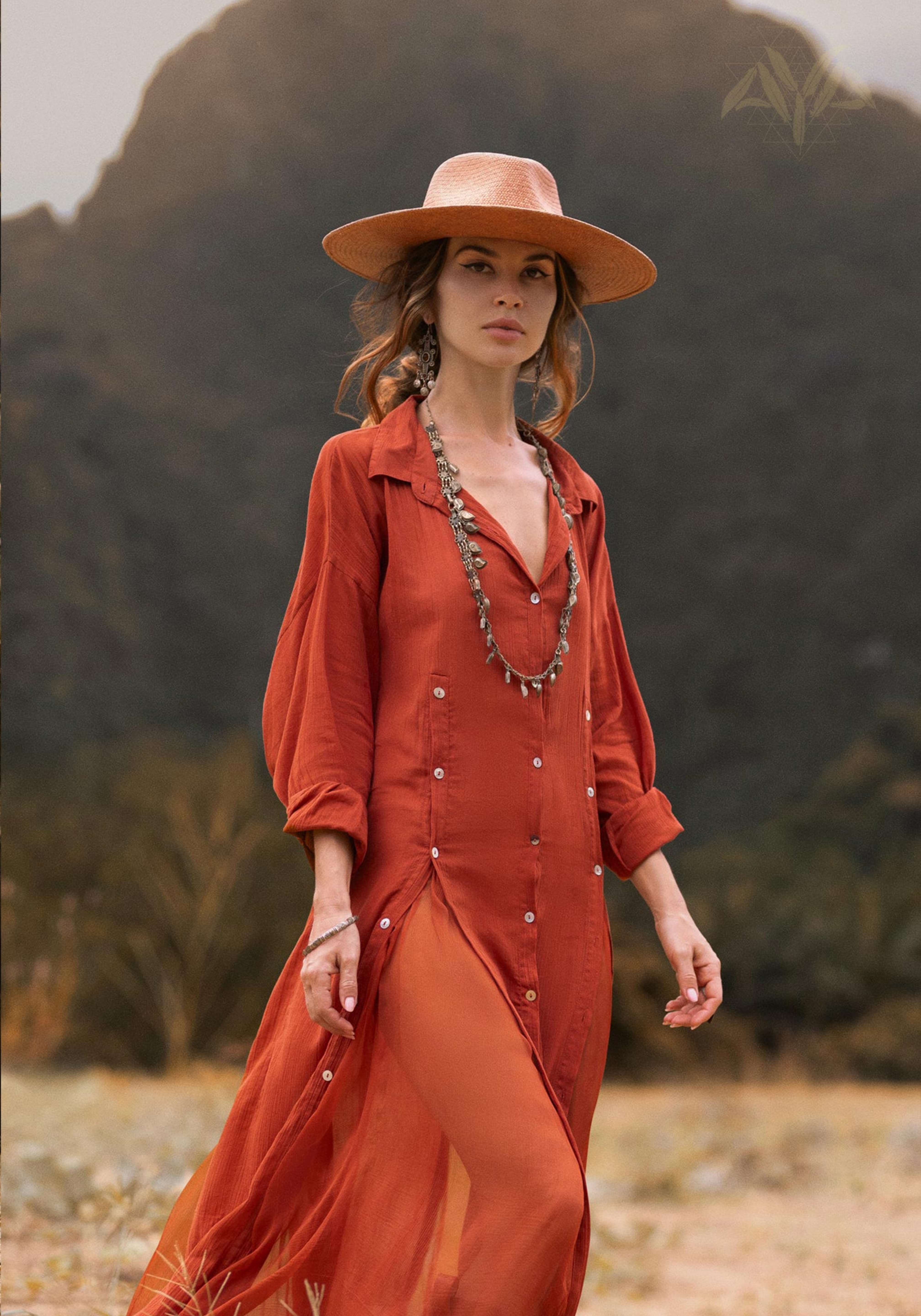 A woman in a flowing Red Kannika Shirt Dress by AYA Sacred Wear strolls through nature against a mountainous backdrop. The adjustable kaftan, made from organic crinkle cotton and enhanced by botanical dyes, is complemented by earthy tones. Her long necklace glints as she looks ahead confidently.