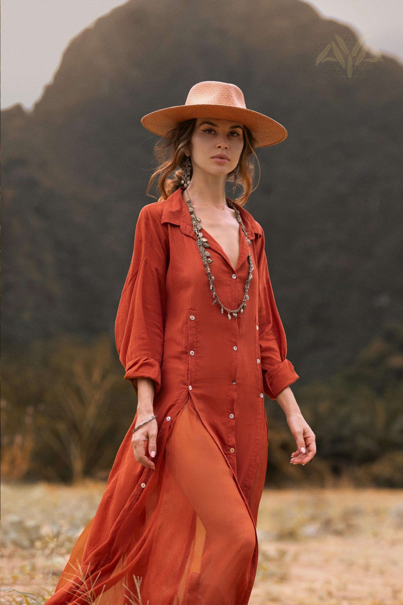 A woman in a flowing Red Kannika Shirt Dress by AYA Sacred Wear strolls through nature against a mountainous backdrop. The adjustable kaftan, made from organic crinkle cotton and enhanced by botanical dyes, is complemented by earthy tones. Her long necklace glints as she looks ahead confidently.