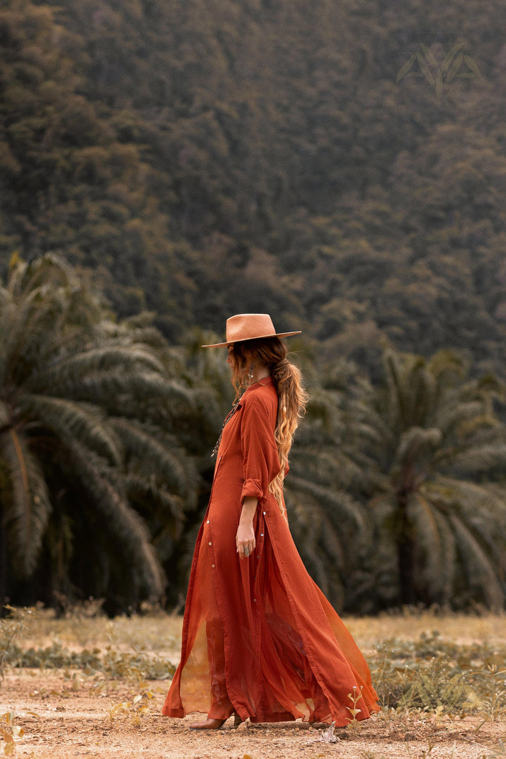 A person wearing a flowing, rust-colored Red Kannika Shirt Dress from AYA Sacred Wear and a wide-brimmed hat stands in an outdoor setting with dense green foliage and palm trees in the background. The person's long hair cascades down their back, enhancing their serene pose and blending harmoniously with the natural environment.