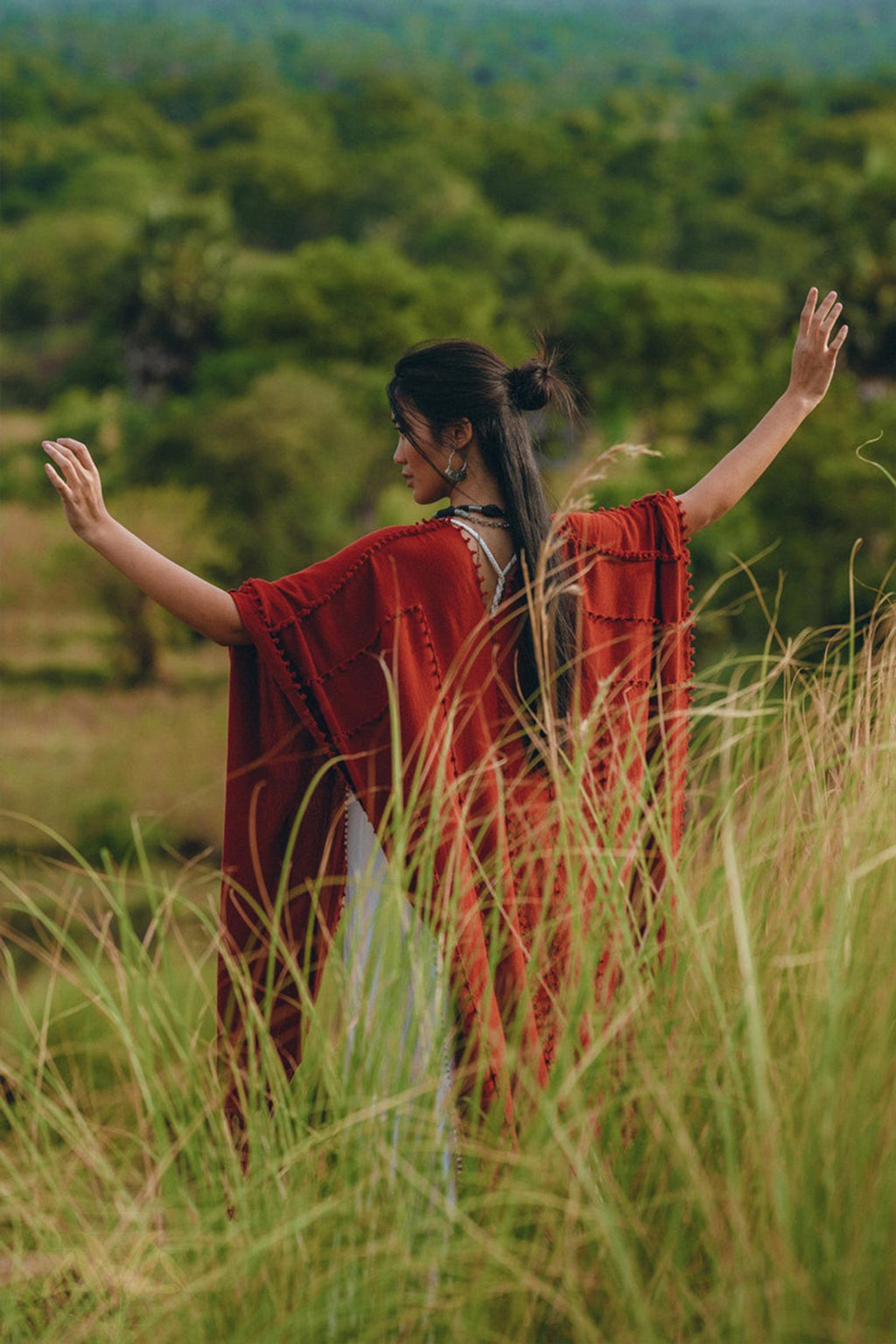 Amidst the lush, green landscape, a person stands with outstretched arms in tall grass. They wear the Red Poncho Robe by AYA Sacred Wear, a flowing bohemian garment that resembles a poncho cape. Their long dark hair is tied up, complementing the essence of traditional crafts that Bali is renowned for.