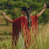 Amidst the lush, green landscape, a person stands with outstretched arms in tall grass. They wear the Red Poncho Robe by AYA Sacred Wear, a flowing bohemian garment that resembles a poncho cape. Their long dark hair is tied up, complementing the essence of traditional crafts that Bali is renowned for.