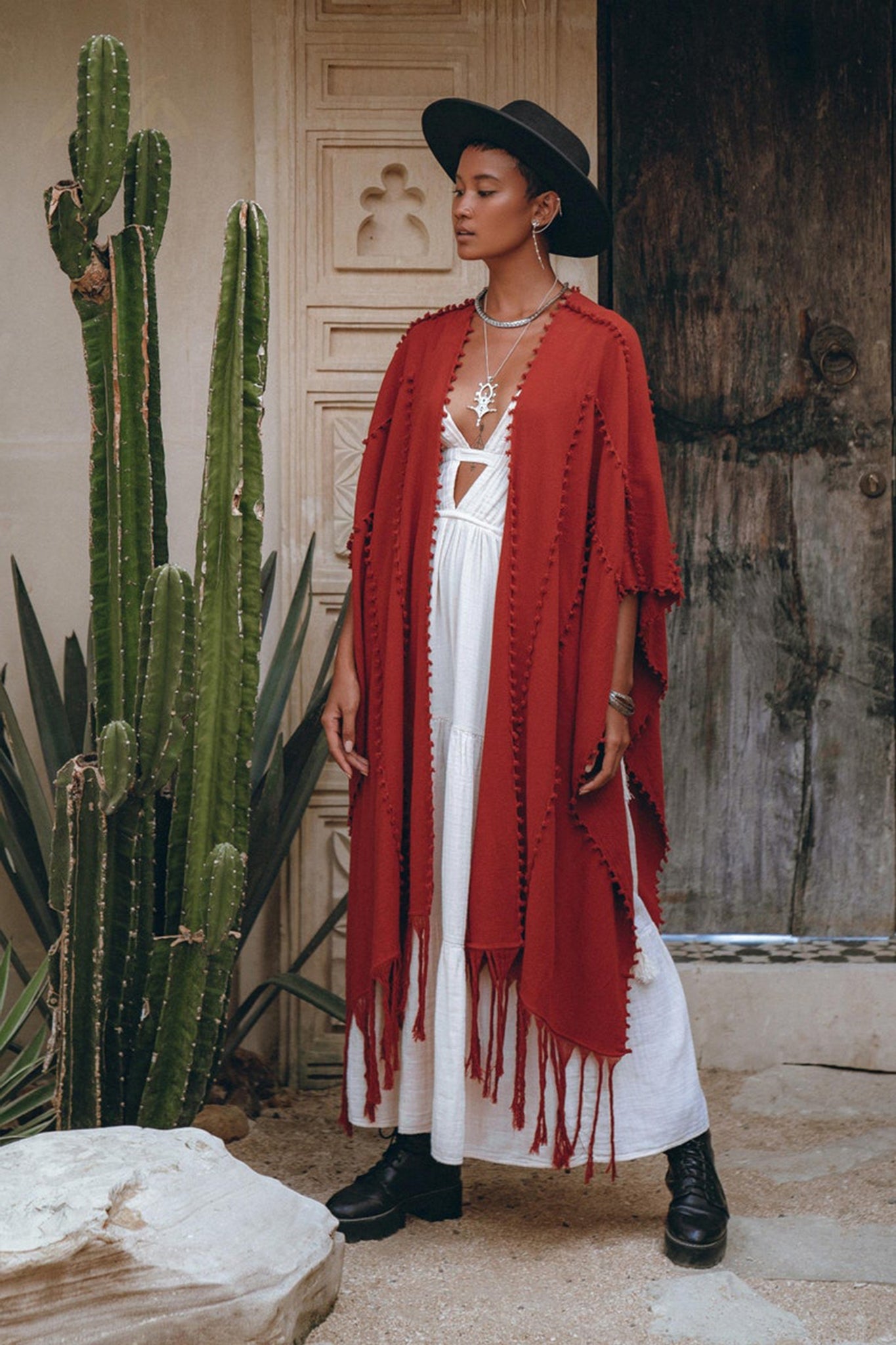 A person draped in a Red Poncho Robe from AYA Sacred Wear's Boho Cape collection for women pairs it with a flowing white dress while posing beside a tall cactus in a rustic setting. Their ensemble is completed with a black wide-brimmed hat, boots, and an intricately designed necklace. The scene is set against the backdrop of a wooden door and textured wall.