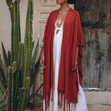 A person draped in a Red Poncho Robe from AYA Sacred Wear's Boho Cape collection for women pairs it with a flowing white dress while posing beside a tall cactus in a rustic setting. Their ensemble is completed with a black wide-brimmed hat, boots, and an intricately designed necklace. The scene is set against the backdrop of a wooden door and textured wall.