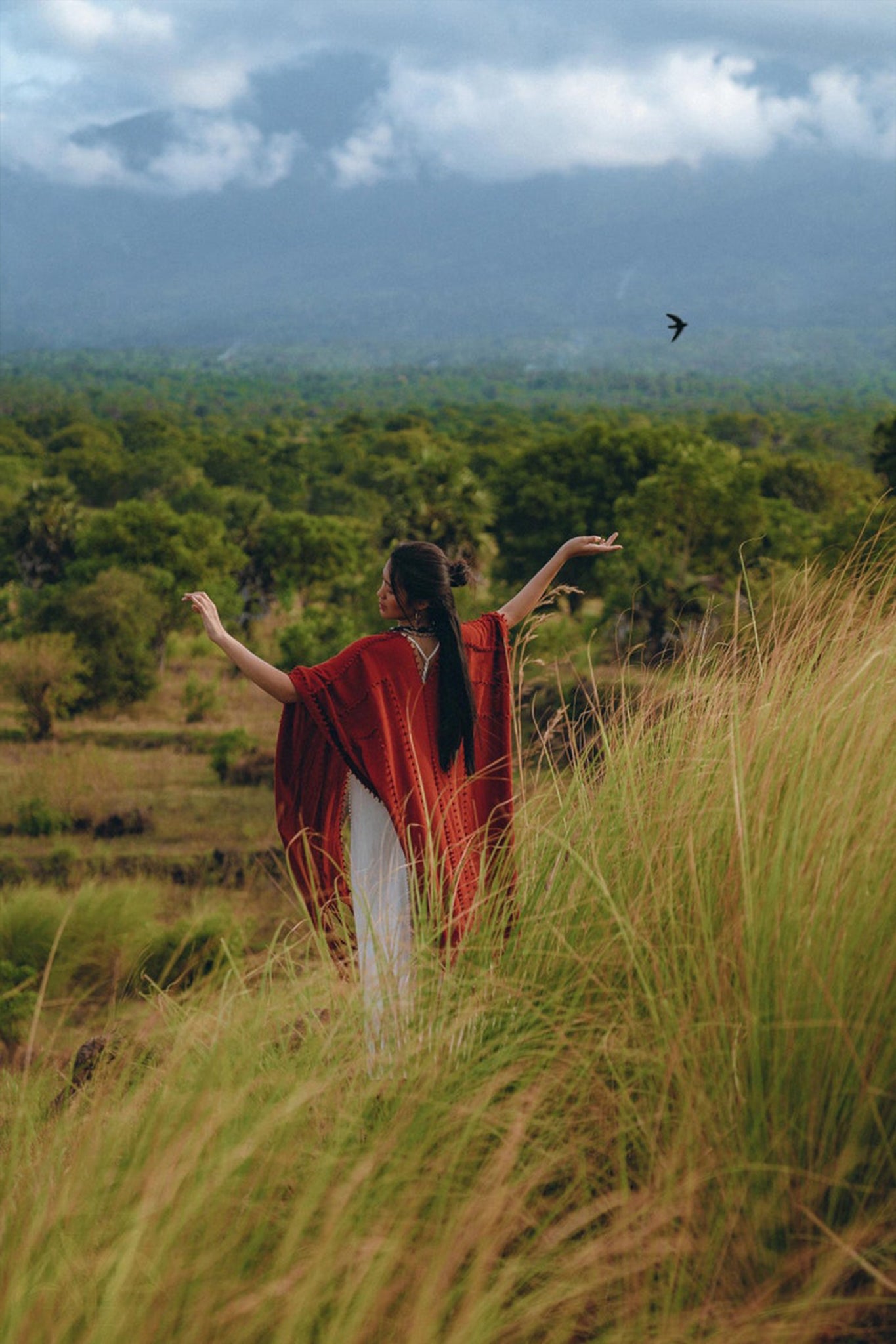 A person wearing an AYA Sacred Wear Red Poncho Robe, a Bohemian Kimono Overcoat with long black hair, stands in tall grass, arms outstretched, facing a lush green landscape with mountains in the background. A bird flies nearby under a cloudy sky, capturing the essence of traditional crafts from Bali.