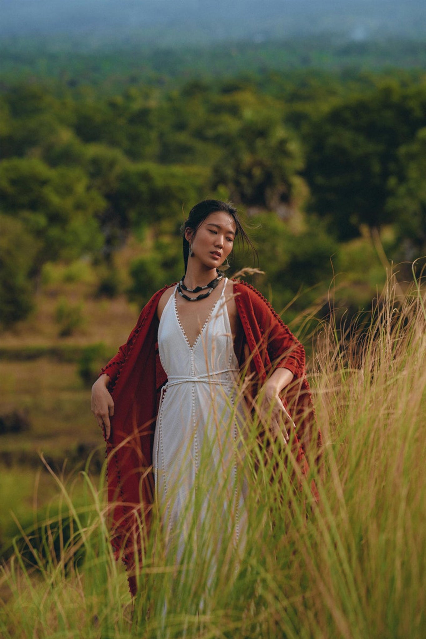 A person dressed in a white dress and an AYA Sacred Wear red Boho Cape, known as the Poncho Robe or Bohemian Kimono Overcoat, stands in a field of tall grass. The scene is set against lush greenery and hills under a cloudy sky.