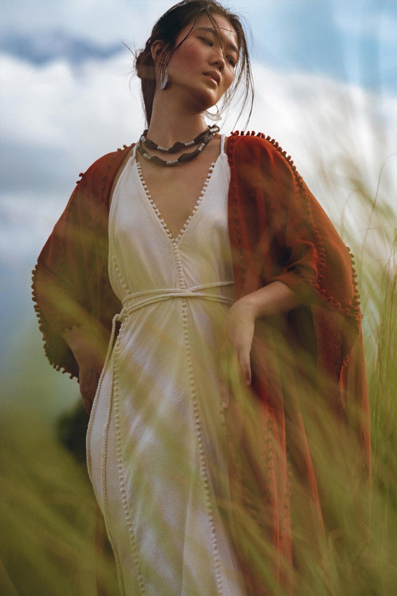 A woman wearing a white dress and the Red Poncho Robe by AYA Sacred Wear stands elegantly in a field of tall grass, under a cloudy sky. Her serene expression complements her loosely tied-back hair.