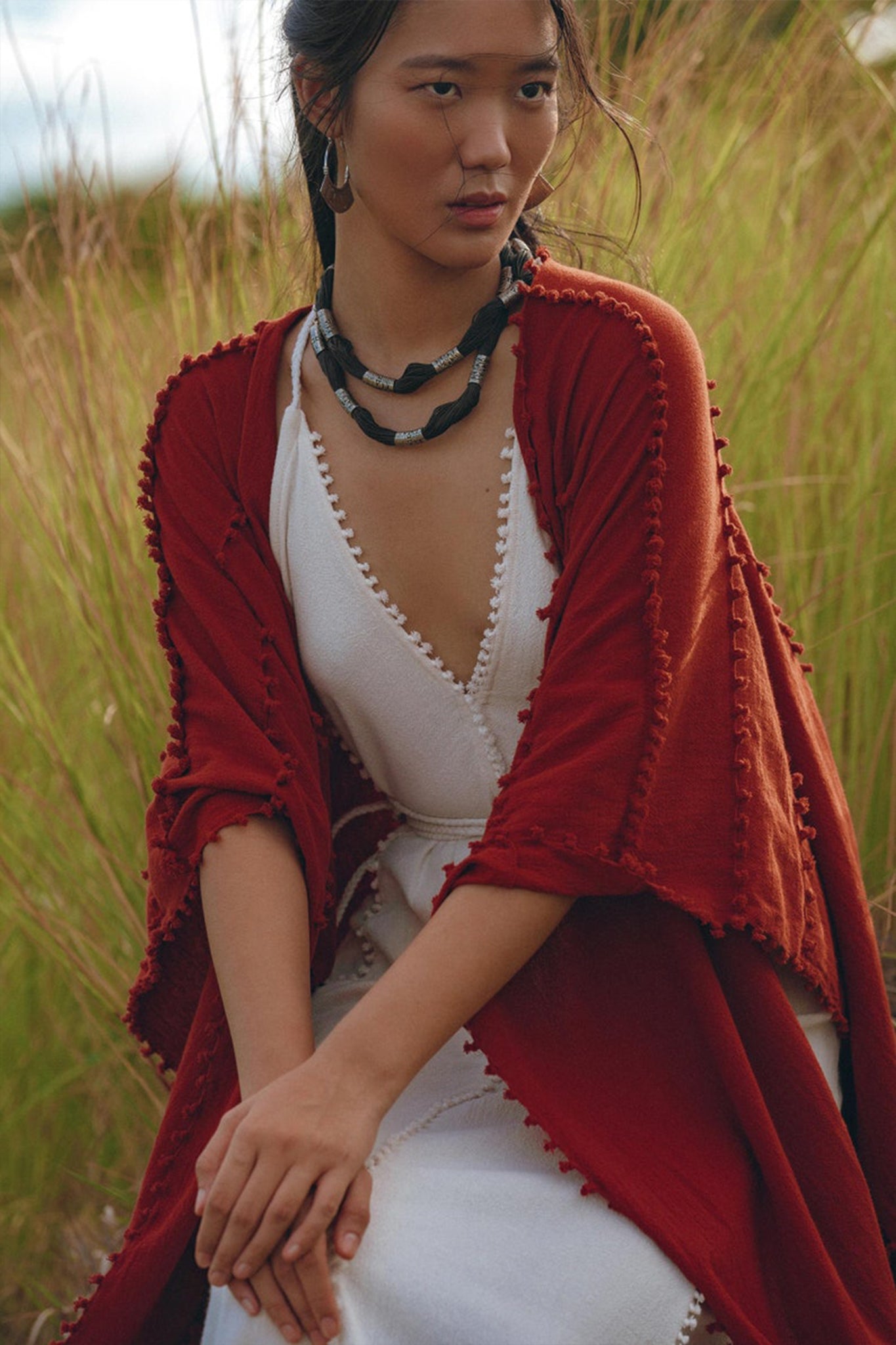 A person dressed in white and wrapped in a rust-colored Red Poncho Robe from AYA Sacred Wear sits in a grassy field. They are accessorized with a black beaded necklace and hoop earrings, creating an earthy, natural vibe that resonates with the charm of traditional Balinese crafts.