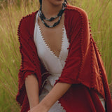 A person dressed in white and wrapped in a rust-colored Red Poncho Robe from AYA Sacred Wear sits in a grassy field. They are accessorized with a black beaded necklace and hoop earrings, creating an earthy, natural vibe that resonates with the charm of traditional Balinese crafts.