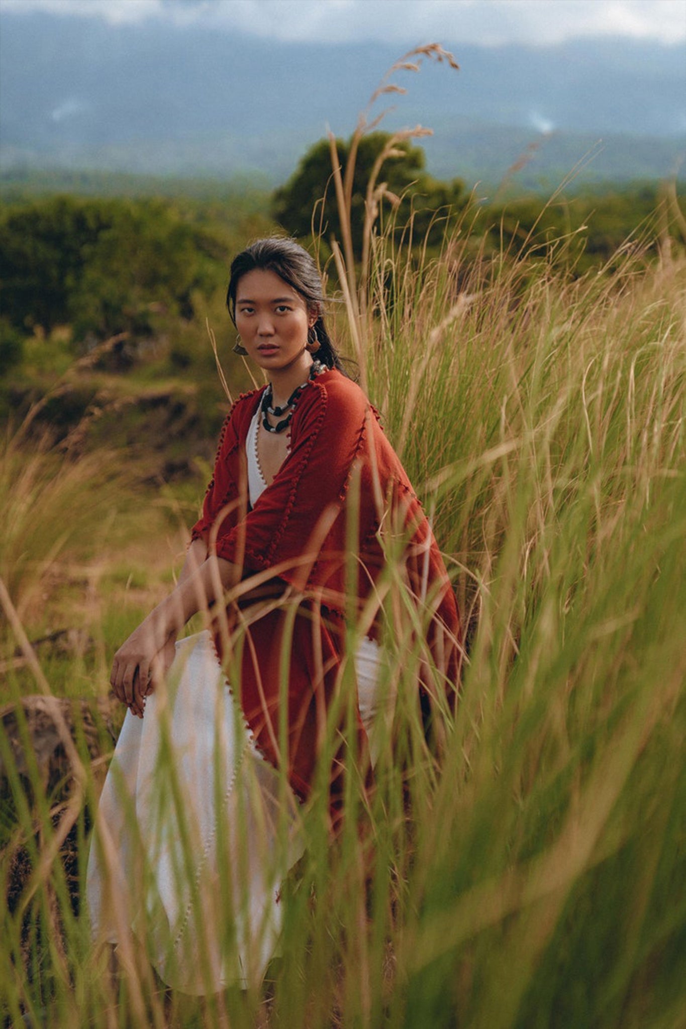 Draped in an AYA Sacred Wear Red Poncho Robe, a woman wearing a Boho Cape and white dress sits amidst tall grass in a serene, lush landscape with hills in the background. The atmosphere is calm and contemplative, evoking a connection to traditional crafts.