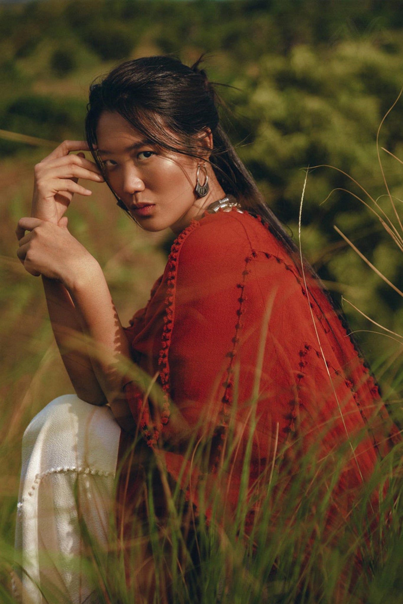 A person wearing a Red Poncho Robe by AYA Sacred Wear, along with white pants, sits gracefully among tall grass. With a focused expression and hand near their face, the lush green background enhances the serene outdoor setting, reminiscent of traditional crafts.