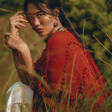 A person wearing a Red Poncho Robe by AYA Sacred Wear, along with white pants, sits gracefully among tall grass. With a focused expression and hand near their face, the lush green background enhances the serene outdoor setting, reminiscent of traditional crafts.