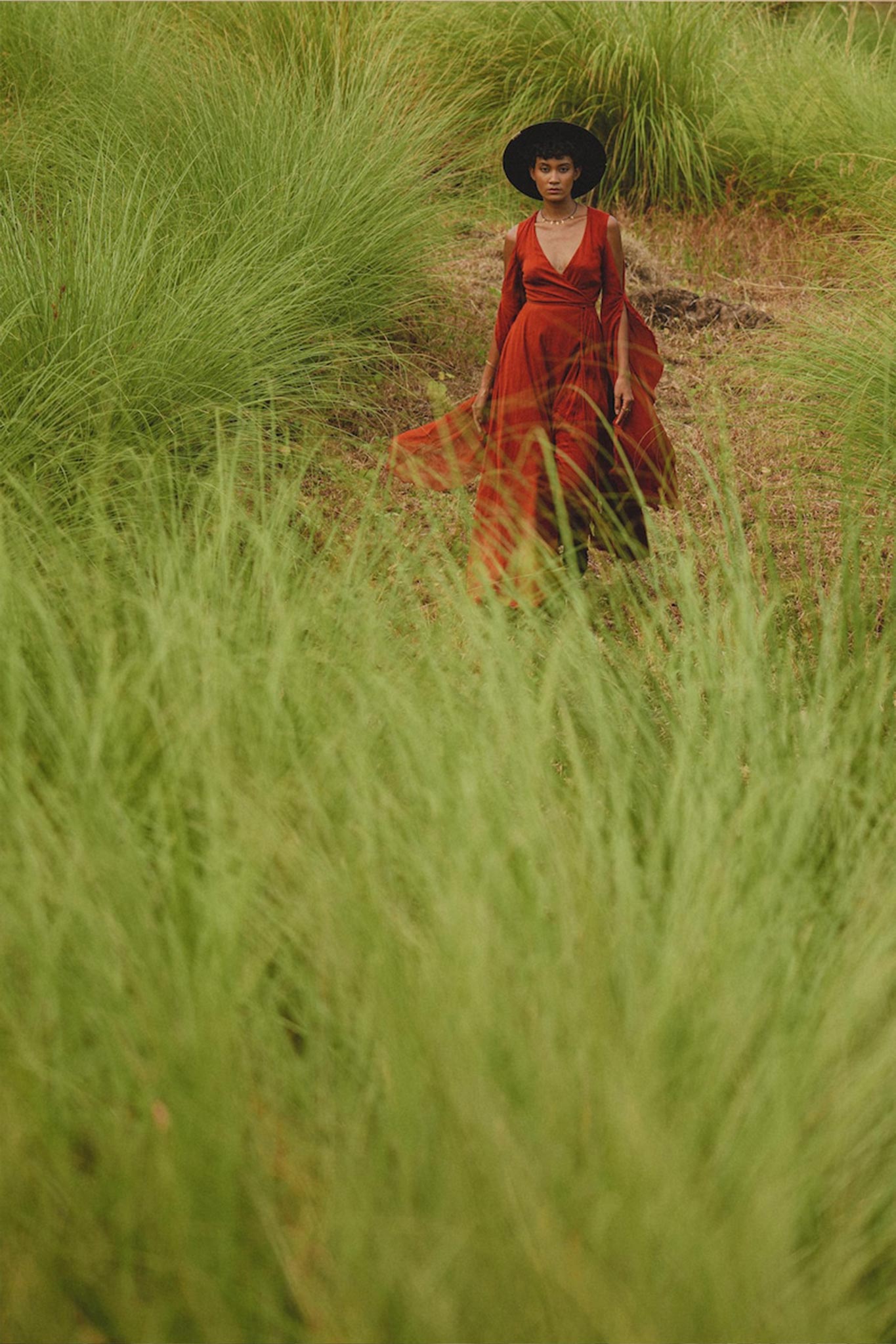 Dressed in the AYA Sacred Wear Red Rose Makosh Goddess Dress, a person glides through tall green grass, their black hat providing an exquisite contrast to the vibrant surroundings.