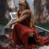 A woman in an AYA Sacred Wear Red Aurora Greek Goddess Dress • Boho Macrame Dress • Boho Sustainable Dress is sitting on a wooden boat covered with pink flowers, surrounded by water, and flanked by tall, rocky cliffs. She has long, wavy hair and her head is tilted back as she poses with her eyes closed.