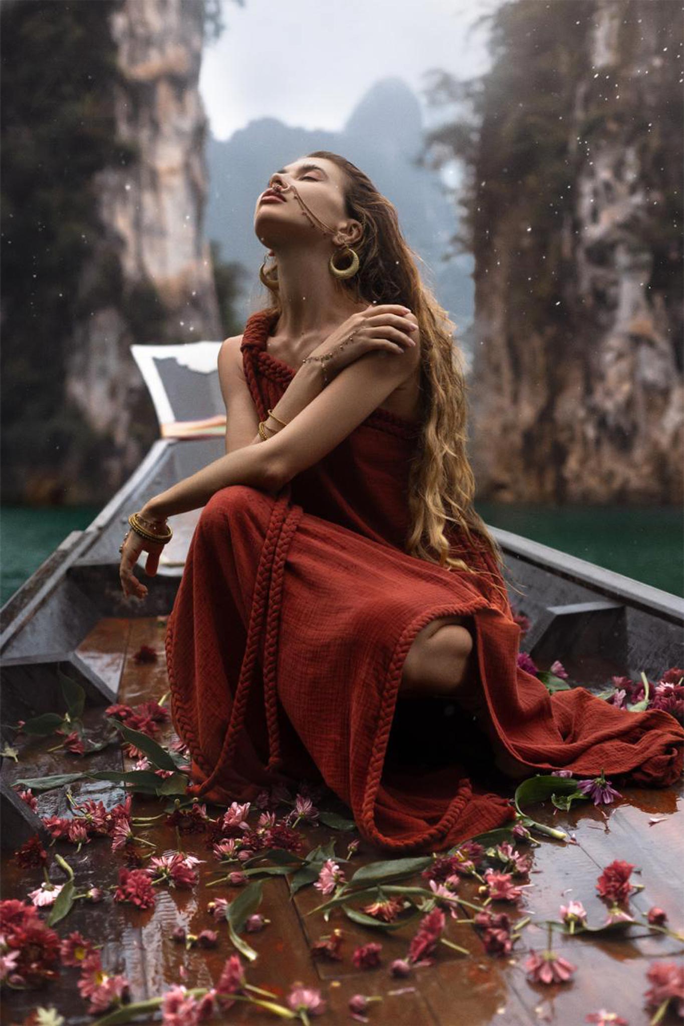 A woman in an AYA Sacred Wear Red Aurora Greek Goddess Dress • Boho Macrame Dress • Boho Sustainable Dress is sitting on a wooden boat covered with pink flowers, surrounded by water, and flanked by tall, rocky cliffs. She has long, wavy hair and her head is tilted back as she poses with her eyes closed.