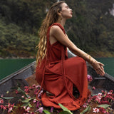 A woman with long, wavy hair wearing an AYA Sacred Wear Red Aurora Greek Goddess Dress • Boho Macrame Dress • Boho Sustainable Dress sits at the end of a wooden boat. She is surrounded by scattered flowers and greenery on the boat's deck. The background features lush green foliage and the calm water of a lake or river.
