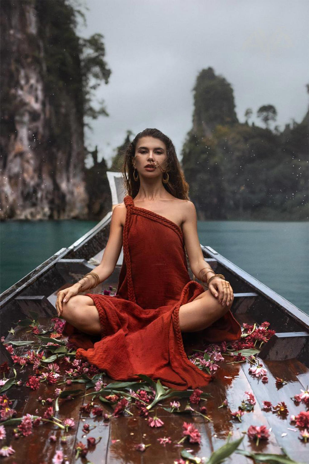 A person wearing an AYA Sacred Wear Red Aurora Greek Goddess Dress • Boho Macrame Dress • Boho Sustainable Dress sits cross-legged at the front of a wooden boat filled with scattered flowers. The boat is on a body of water surrounded by tall, rocky cliffs and lush greenery. The scene is serene, with a misty, overcast sky.