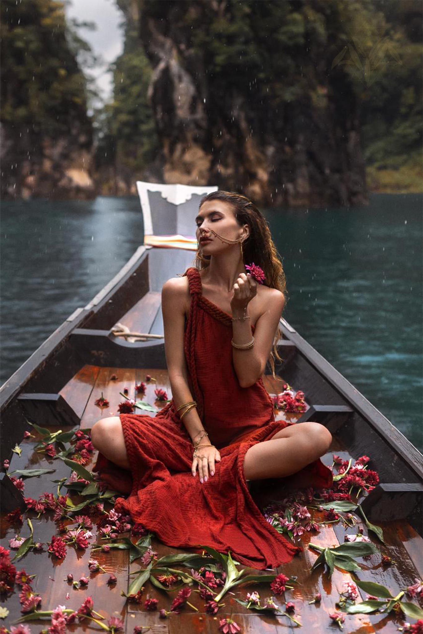 A woman in an AYA Sacred Wear Red Aurora Greek Goddess Dress, also known as the Boho Macrame Dress or Boho Sustainable Dress, sits cross-legged at the front of a wooden boat adorned with pink flowers. She holds a flower close to her face and is surrounded by serene turquoise water with lush, rocky cliffs in the background.
