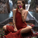 A woman wearing an AYA Sacred Wear Red Aurora Greek Goddess Dress • Boho Macrame Dress • Boho Sustainable Dress sits on a boat surrounded by scattered pink flowers. She holds a purple flower to her mouth, gazing thoughtfully to the side. The boat floats in a serene waterway, with tall rocky formations and lush greenery in the background.