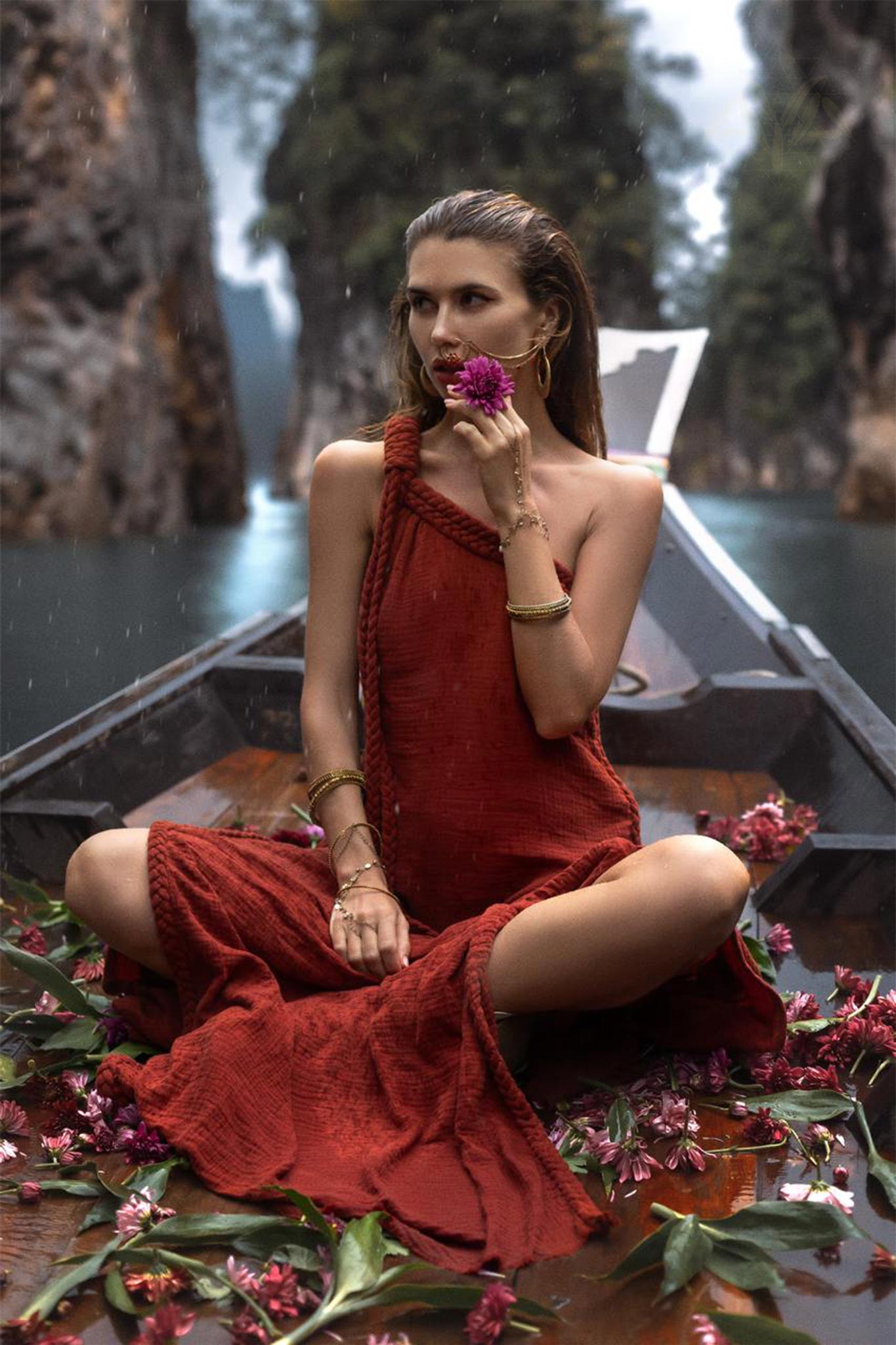 A woman wearing an AYA Sacred Wear Red Aurora Greek Goddess Dress • Boho Macrame Dress • Boho Sustainable Dress sits on a boat surrounded by scattered pink flowers. She holds a purple flower to her mouth, gazing thoughtfully to the side. The boat floats in a serene waterway, with tall rocky formations and lush greenery in the background.