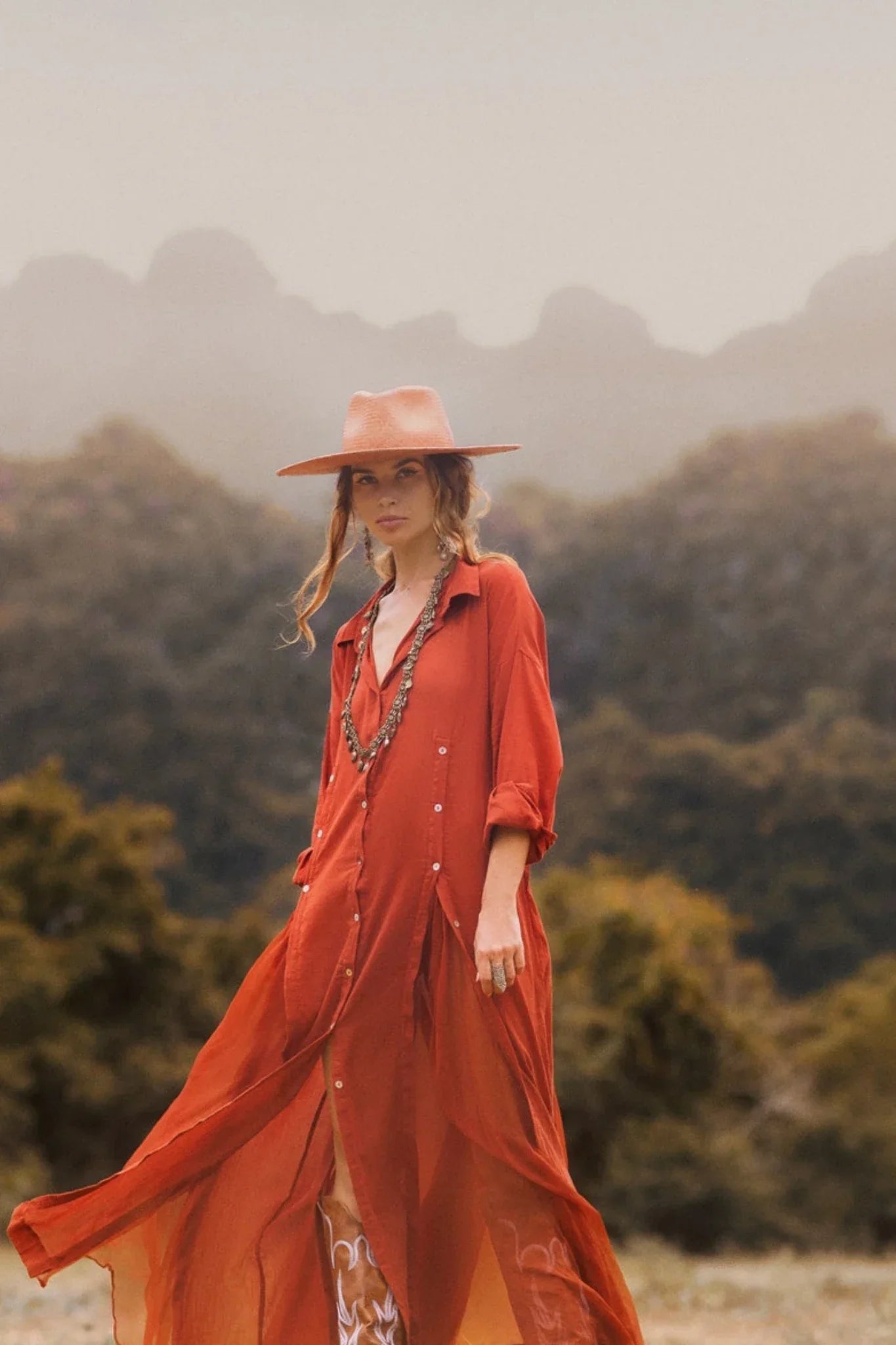 A woman outdoors wears the AYA Sacred Wear Red Kannika Shirt Dress—an adjustable, multiway button-down kaftan—paired with a wide red hat and layered jewelry. Misty trees and mountains blur in the background.
