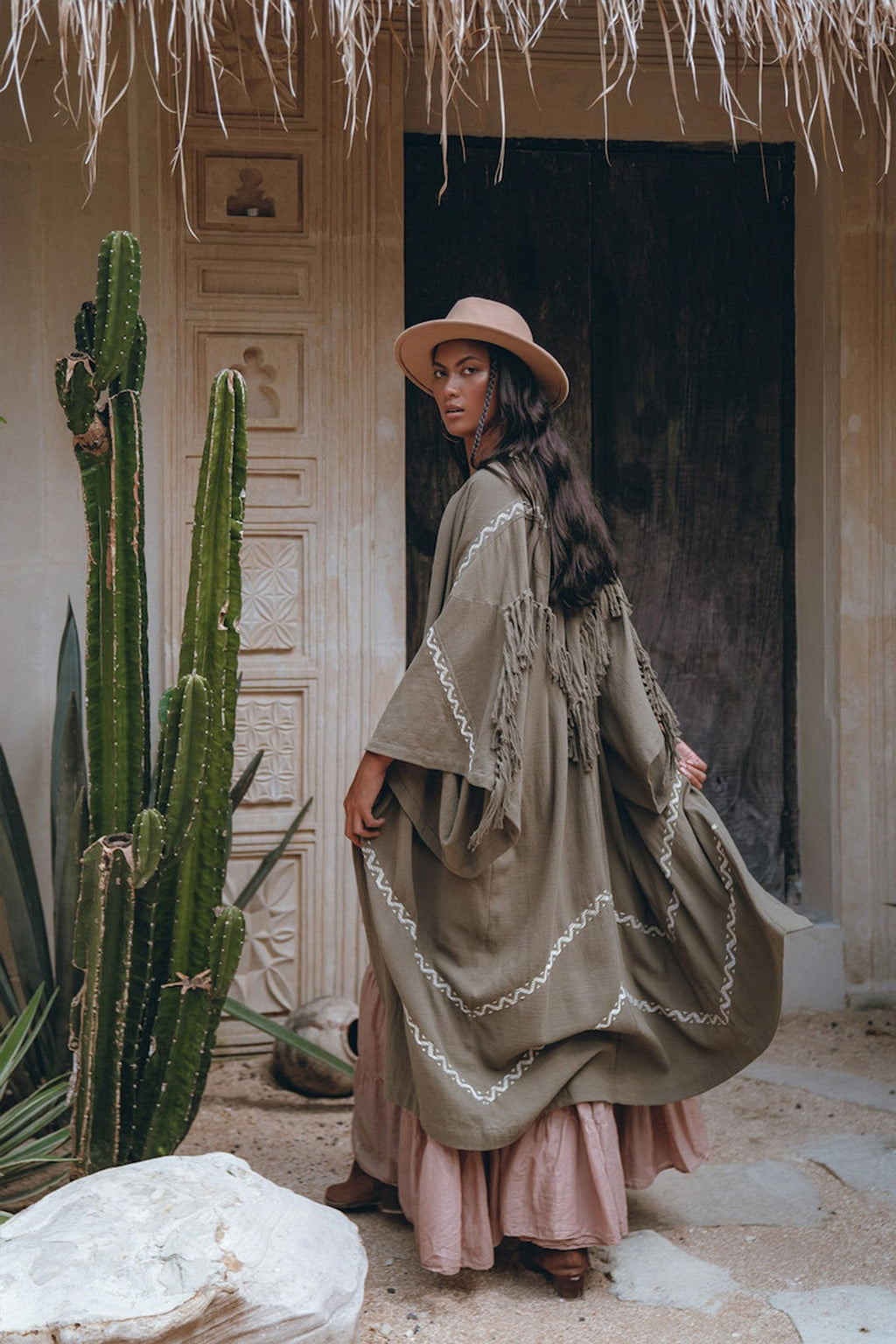 A person wearing the AYA Sacred Wear Sage Colour Organic Cotton Poncho Cape, featuring a bohemian design, stands beside tall cacti in front of a richly carved wooden door. The setting exudes a rustic and bohemian vibe.