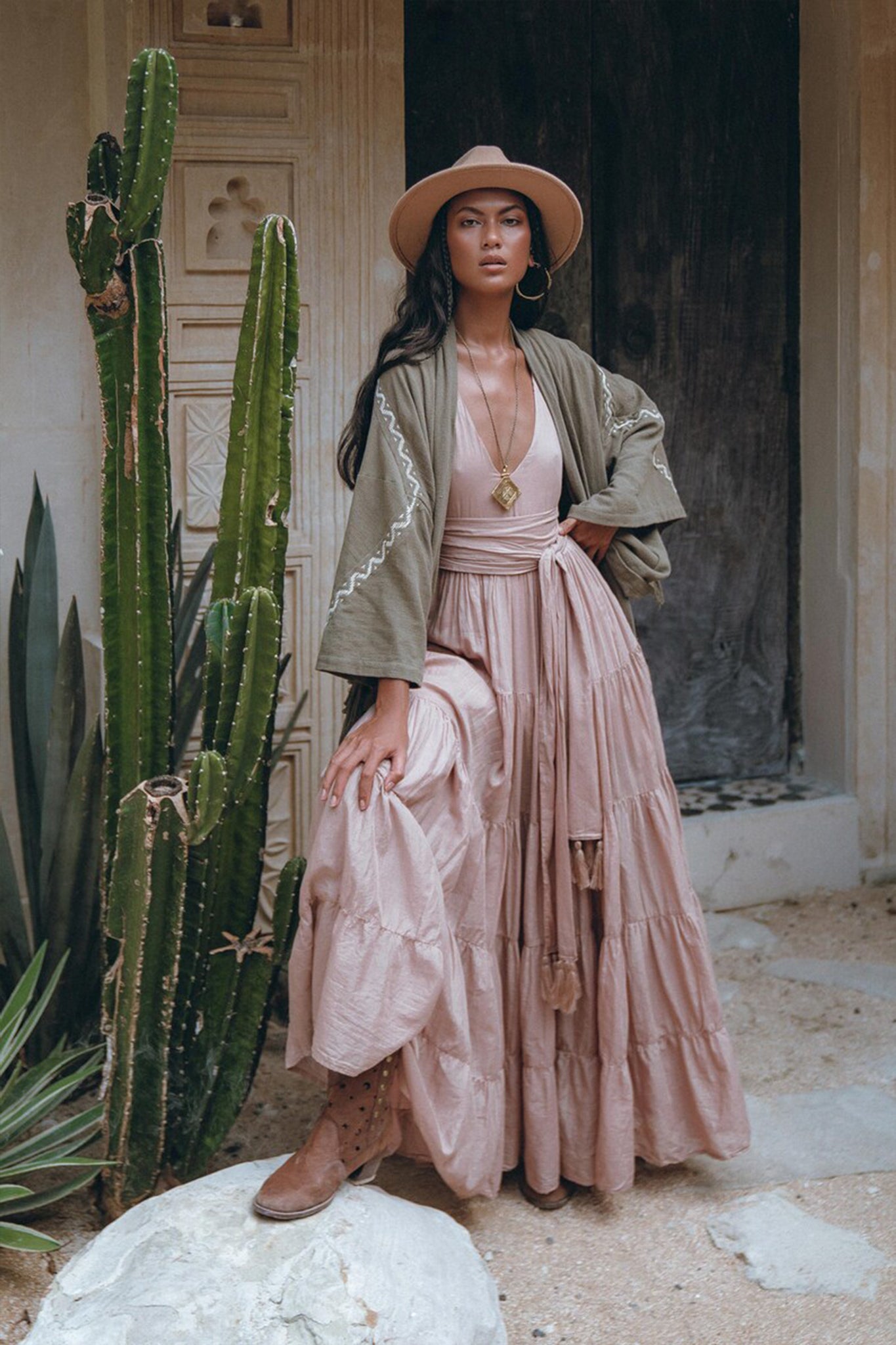 A woman wearing a wide-brimmed hat and pink maxi dress stands beside a towering cactus. She confidently poses in front of a rustic wooden door in the desert, draped in an AYA Sacred Wear Sage Colour Organic Cotton Poncho Cape, paired with brown boots.