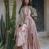 A woman wearing a wide-brimmed hat and pink maxi dress stands beside a towering cactus. She confidently poses in front of a rustic wooden door in the desert, draped in an AYA Sacred Wear Sage Colour Organic Cotton Poncho Cape, paired with brown boots.