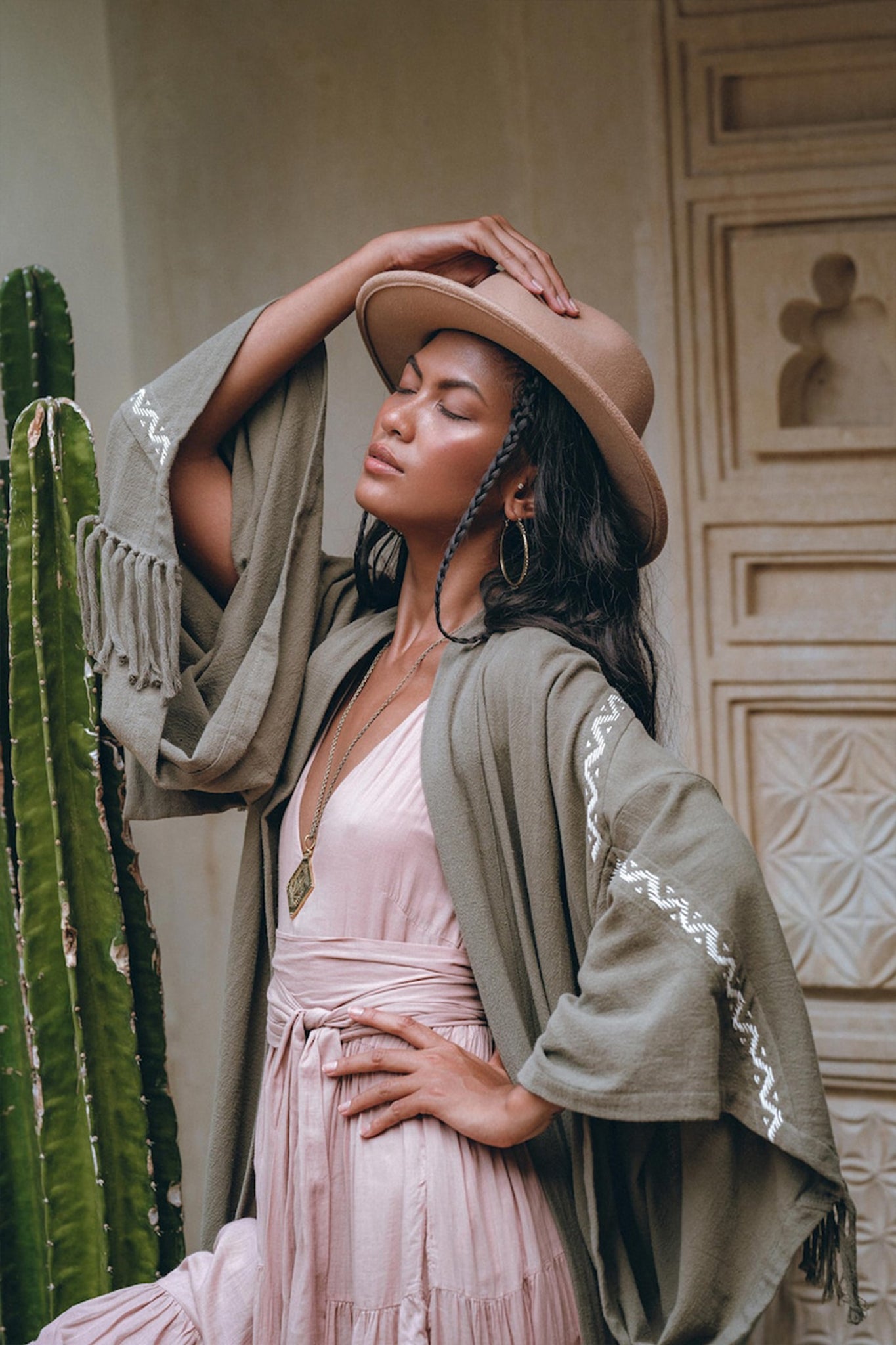 A woman wearing the Sage Colour Organic Cotton Poncho Cape by AYA Sacred Wear stands serenely with her eyes closed. She is dressed in a pink dress adorned with shamanic embroidery, complemented by a tan hat and large hoop earrings. Her green shawl, draped over the bohemian cardigan, adds to her tranquil aura as she leans against a tall cactus in a rustic setting with intricately carved wooden doors, gently placing one hand on her hat.