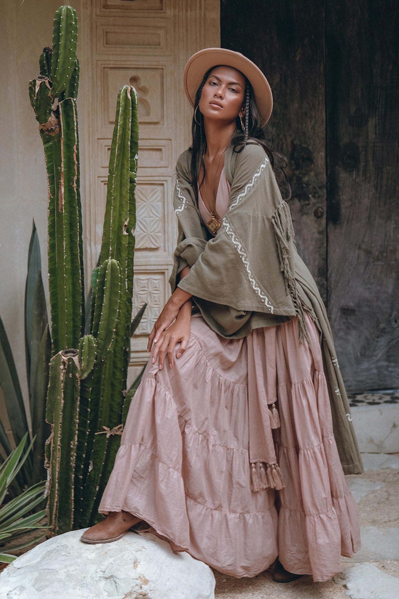 A woman wearing an AYA Sacred Wear Sage Colour Organic Cotton Poncho Cape, styled as a bohemian cardigan, poses beside tall cacti against an ornate door. She is dressed in a wide-brimmed hat, a layered dress with shamanic embroidery patterns, and brown boots, surrounded by a rustic, earthy setting.