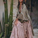 A woman wearing an AYA Sacred Wear Sage Colour Organic Cotton Poncho Cape, styled as a bohemian cardigan, poses beside tall cacti against an ornate door. She is dressed in a wide-brimmed hat, a layered dress with shamanic embroidery patterns, and brown boots, surrounded by a rustic, earthy setting.