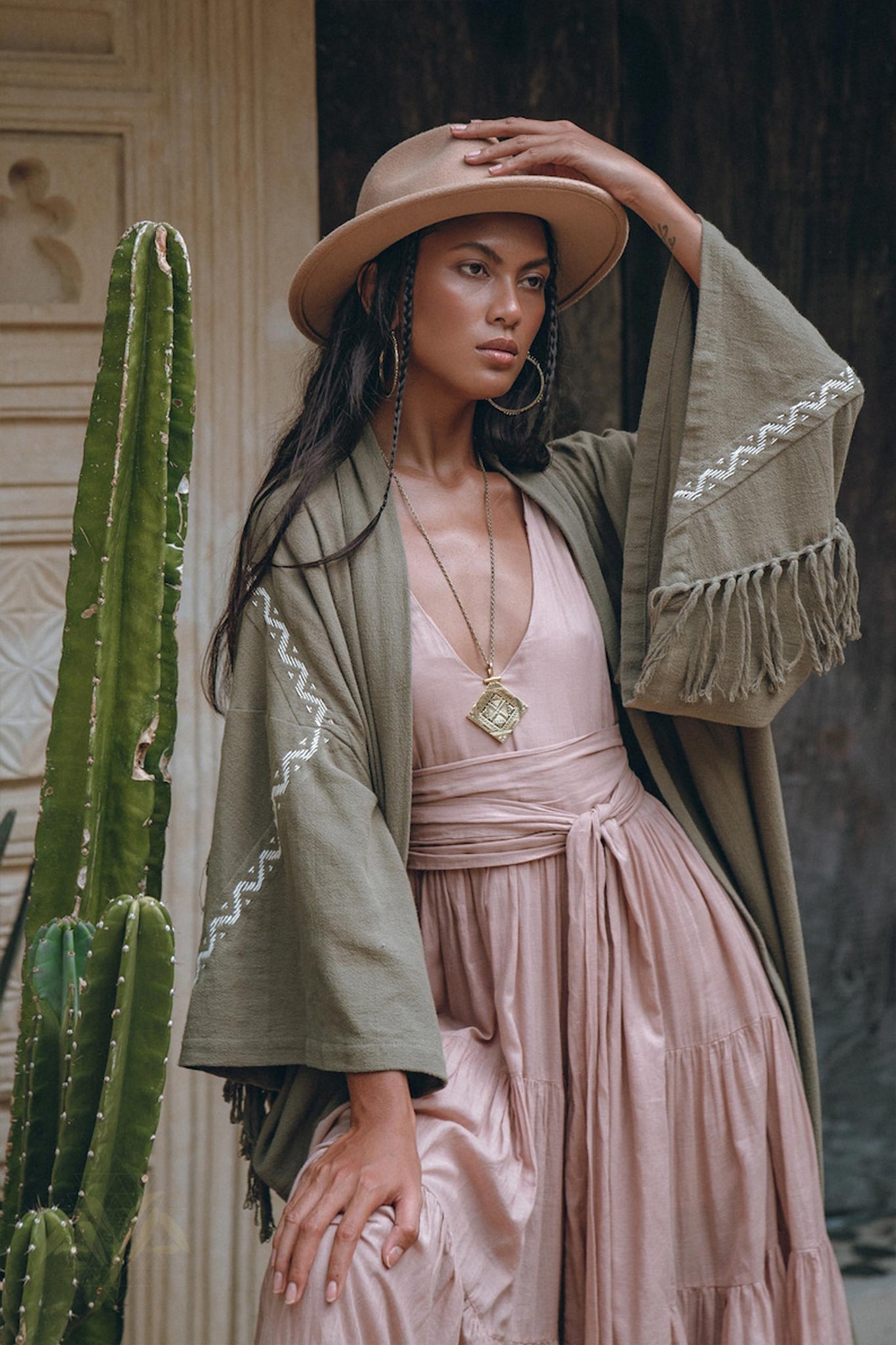 A woman poses confidently in a flowing pink dress, wrapped in the Sage Colour Organic Cotton Poncho Cape by AYA Sacred Wear, featuring shamanic embroidery. She accessorizes with a wide-brimmed tan hat and a pendant necklace. A tall cactus stands beside her against a textured wall backdrop.