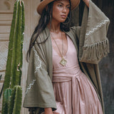 A woman poses confidently in a flowing pink dress, wrapped in the Sage Colour Organic Cotton Poncho Cape by AYA Sacred Wear, featuring shamanic embroidery. She accessorizes with a wide-brimmed tan hat and a pendant necklace. A tall cactus stands beside her against a textured wall backdrop.