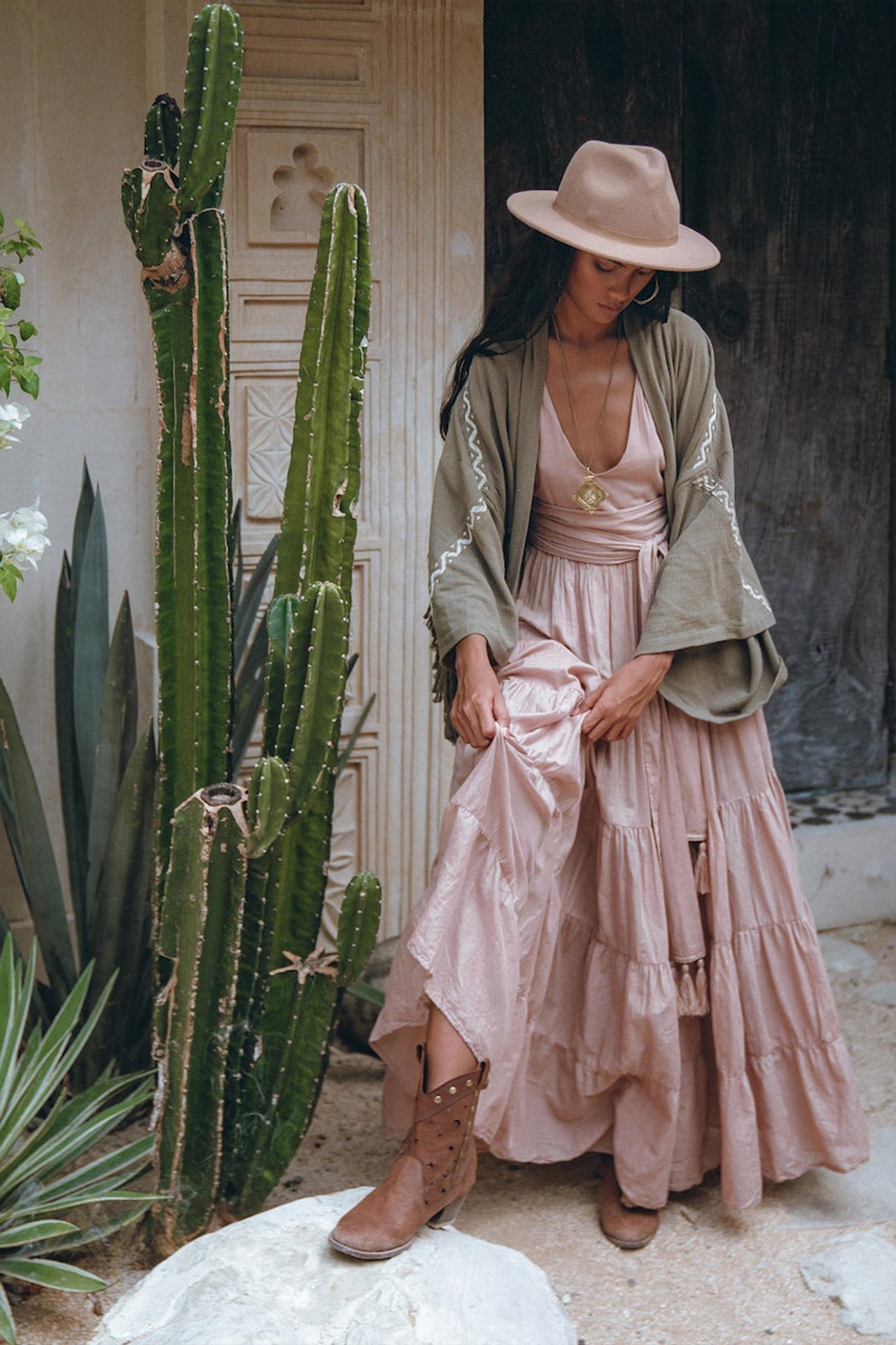 A woman stands beside a tall cactus, gazing downward, adorned in an AYA Sacred Wear ensemble: a tan hat and Sage Colour Organic Cotton Poncho Cape with shamanic embroidery. Her flowing pink dress is complemented by a pendant and brown boots, perfectly capturing her bohemian style.